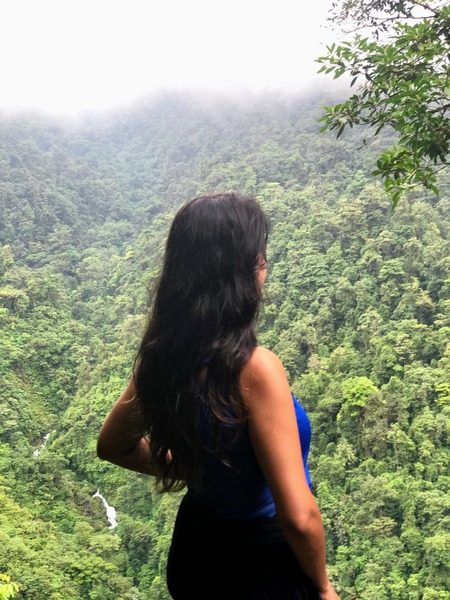 woman with sleeveless blue tank top and black pants, with long black hair standing looking out over a valley of green treetops