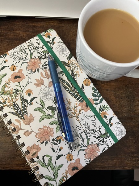 white spiral-bound journal with pink and green floral print, a blue pen set upon it and a milky cup of coffee next to it