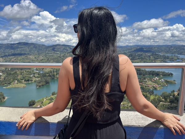 woman in black dress with long black hair with her back to the camera, looking out over blue skies, mountains and a lake