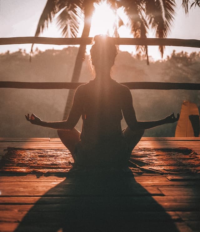 silhouette of woman sitting cross leged with arms resting on knees and meditating outdoors during sunset