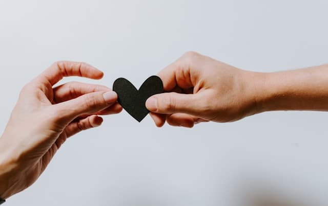 two hands from opposite ends of photo holding a small black paper heart in the middle, one from each side