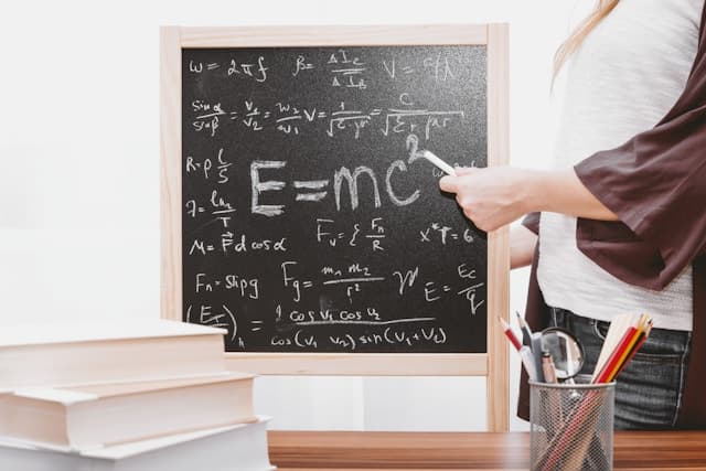 small square blackboard with math formulas on it like E=mc swuared and a hand pointing to it with a piece of white chalk