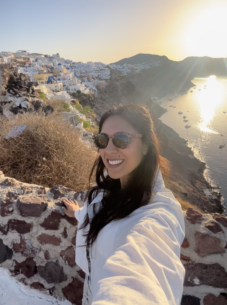 woman in white shirt with dark hair and sunglasses, taking smiling selfie in front of sunrise over Oia, Santorini