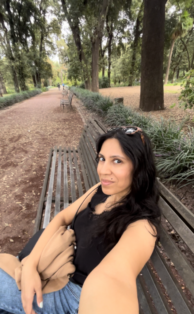 woman in black tank top with black hair and sunglasses on head sitting on a bench taking a selfie of herself smiling